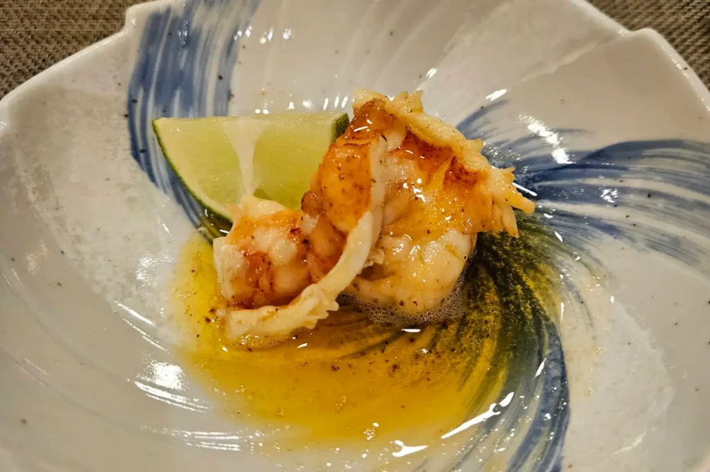 A close-up shot presents a piece of lightly seared lobster or prawn sitting in a rich, buttery yellow sauce with a hint of foam. The seafood is served in a Japanese ceramic bowl painted with deep blue brushstrokes and garnished with a fresh wedge of lime.