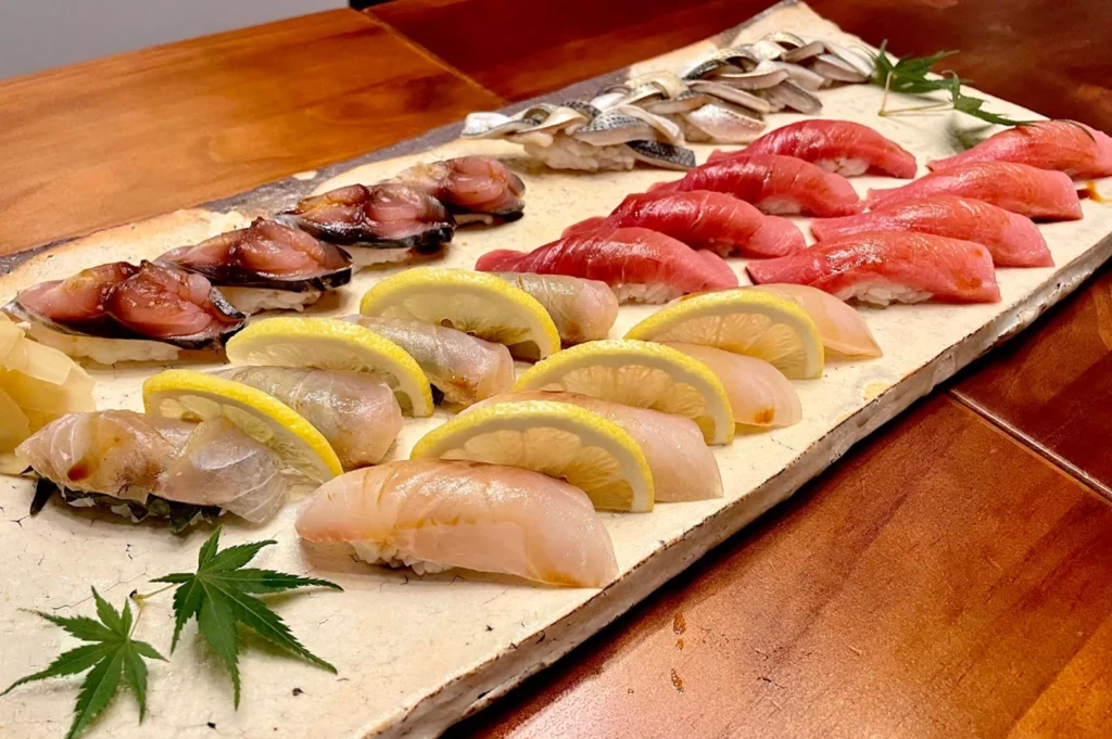 A long, rectangular, rustic cream-colored ceramic plate displays a curated selection of nigiri sushi, many topped with thin, crescent-shaped lemon slices. The pieces are arranged in neat rows on a warm-toned wooden table, with two small, decorative green maple leaves placed at the corners to emphasize a seasonal, artistic presentation.