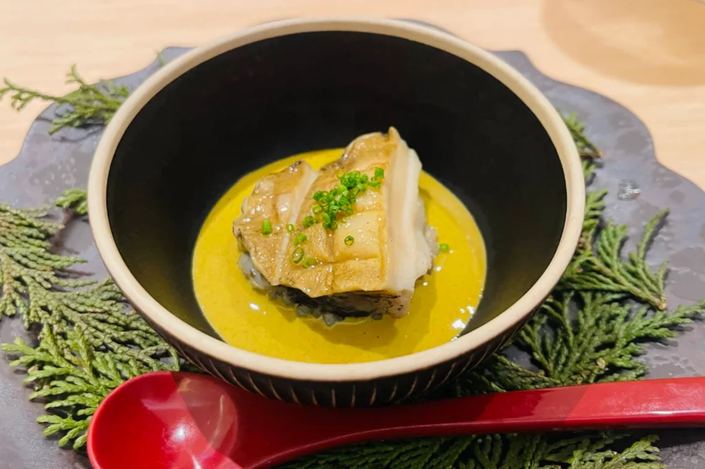 A small, elegant portion of cooked abalone is served in a dark, bowl-like dish filled with a rich, yellow-toned sauce. The abalone is garnished with tiny, vibrant green chopped scallions, and the bowl rests on a dark, rustic plate that is decorated with fresh green evergreen sprigs and accompanied by a sleek, red ceramic spoon.