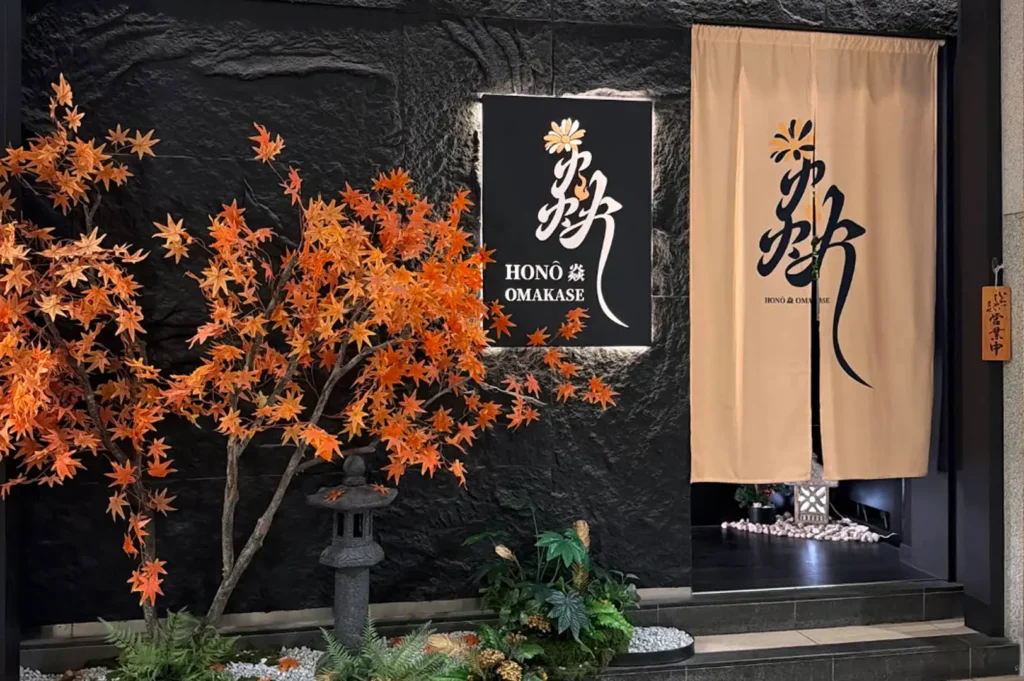 The sleek entrance of Honô Omakase set against textured black stone walls, featuring an illuminated sign displaying Japanese calligraphy, a light beige traditional split doorway curtain (noren), a miniature Japanese stone lantern surrounded by white pebbles, and a decorative artificial maple tree with vibrant orange autumn leaves.