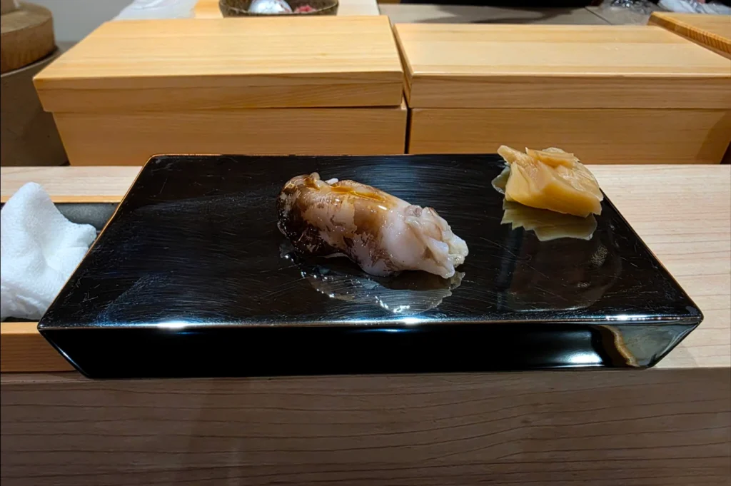 A piece of nigiri sushi with a uniquely textured, glistening topping is presented on a minimalist black rectangular plate, accompanied by a small portion of pickled ginger on the side.