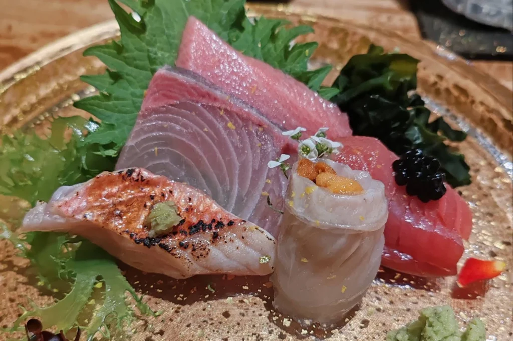 A vibrant close-up displays an assortment of fresh sashimi and specialty seafood arranged on a textured glass plate with gold flecks. The selection features thick, beautifully marbled slices of pink tuna belly (otoro), deep red tuna loin, and sleek silver-skinned fish arranged over fresh green shiso leaves. In the foreground, a piece of lightly seared white fish with a dab of green wasabi sits next to a delicate roll of white fish filled with rich orange sea urchin (uni), all accented by tiny white edible flowers, a mound of dark seaweed, and a dollop of fresh wasabi.
