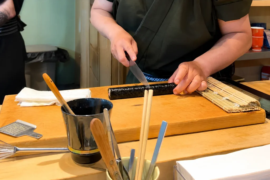 A mid-section shot captures a sushi chef dressed in a traditional dark green uniform using a long, professional knife to slice a freshly rolled nori-wrapped sushi roll on a thick wooden cutting board. The chef's station is arranged with various culinary tools, including a bamboo rolling mat, kitchen tweezers, wooden spoons, chopsticks in a cup, a small metal grater, and a damp white towel resting on the corner of the workspace.