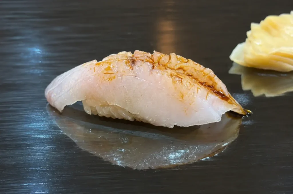 A close-up view of a single piece of delicate white fish nigiri sushi glazed with a light soy sauce coating, resting on a dark, glossy lacquer serving surface with pickled ginger visible in the background.