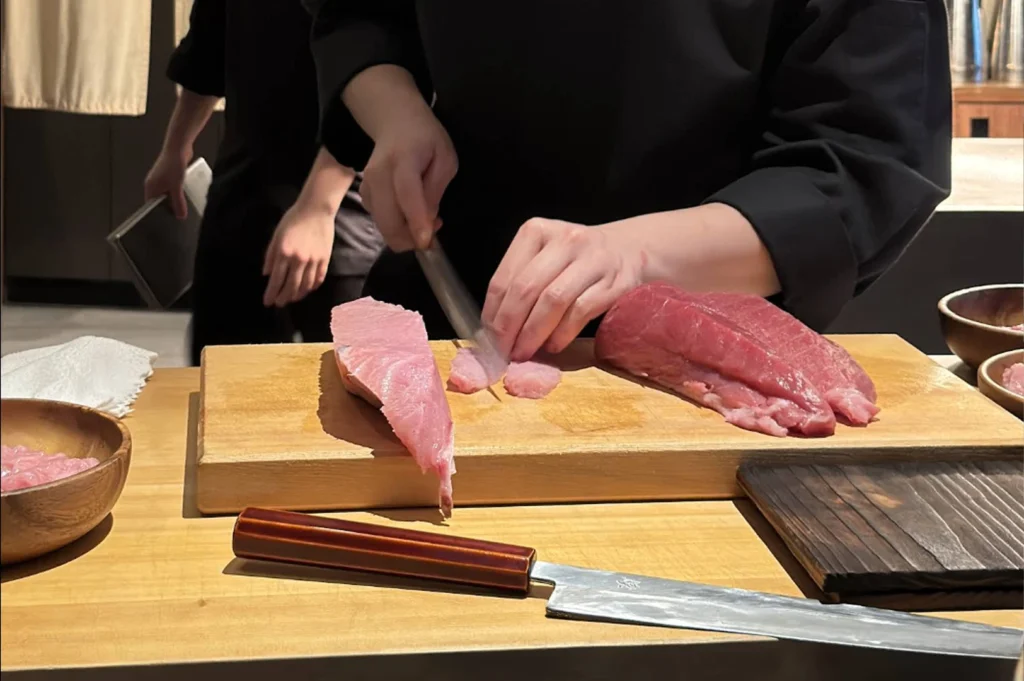 A close-up shot of a chef in a professional black uniform meticulously slicing raw fish on a thick wooden cutting board. Using a long, slender, sharp Japanese sashimi knife, the chef delicately cuts thin slices from a block of pale pink, highly marbled fatty tuna (otoro). Next to it on the board lies a larger, deep red loin of tuna. In the foreground, another traditional wooden-handled knife rests on the counter, while a second kitchen staff member is partially visible in the background.