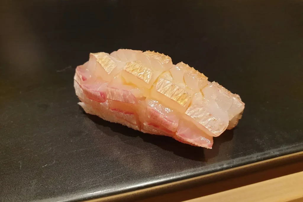 A single piece of nigiri sushi, featuring delicately scored white fish with a light sear on top, is centered on a minimalist, dark gray plate that rests on a clean wooden surface.