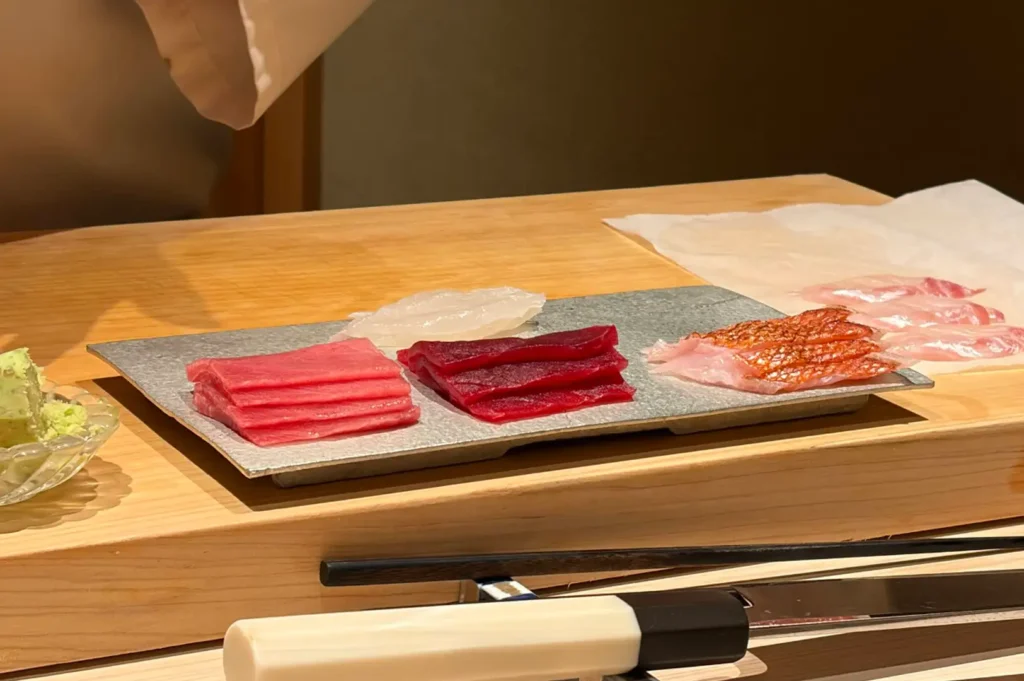A wooden counter setup displaying neatly cut, high-grade sashimi slices—including pink fatty tuna, deep red maguro, translucent white fish, and sea bream with seared skin—laid out on a silver platter next to fresh wasabi and a chef's yanagiba knife.