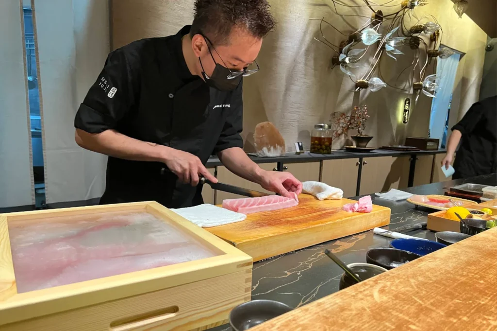 A medium shot captures a male sushi chef wearing glasses and a black face mask, concentrated on slicing a fresh, thick slab of pink-and-white marbled fish on a large wooden cutting board. He wears a professional black chef's jacket with "Sushi Yujo" and his name "Desmond Fong" stitched on it. The marble-textured countertop holds a wooden container filled with ice and fish in the foreground, various small colorful prep bowls, and a decorative modern light fixture resembling glowing glass bulbs hanging against the warm-toned wall in the background.