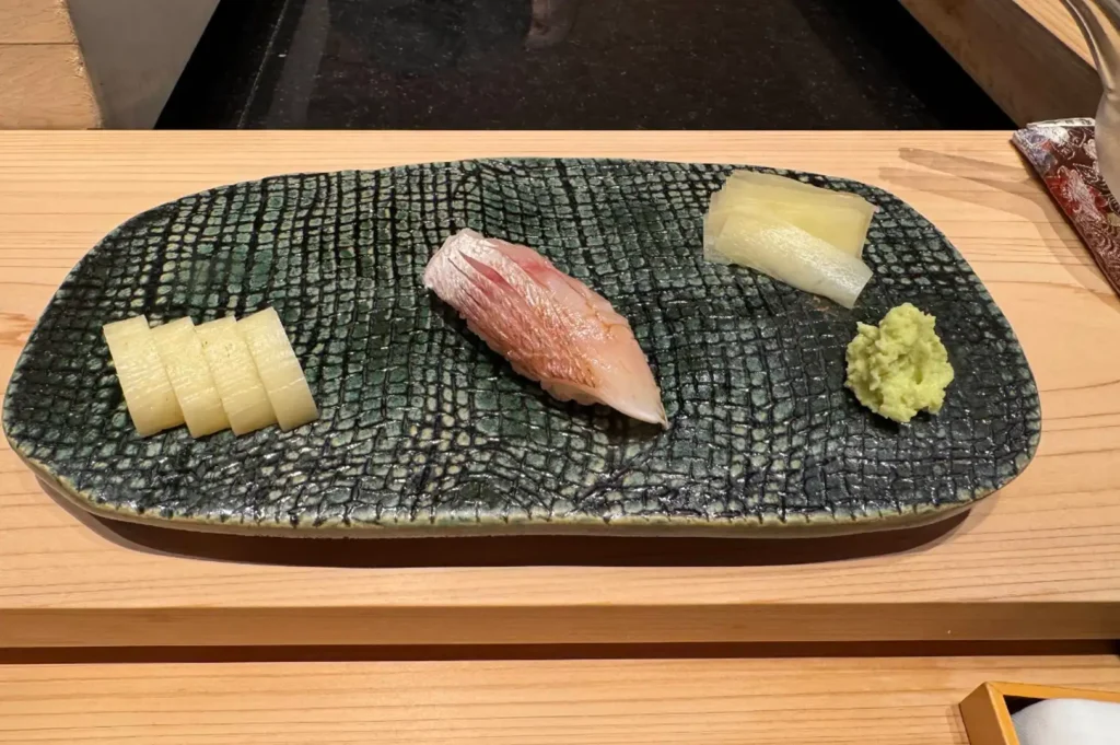 A single piece of nigiri sushi topped with pale pink fish rests on a textured dark green ceramic serving platter on a light wooden table, accompanied by sliced pickled radish, ginger, and a fresh dollop of green wasabi.