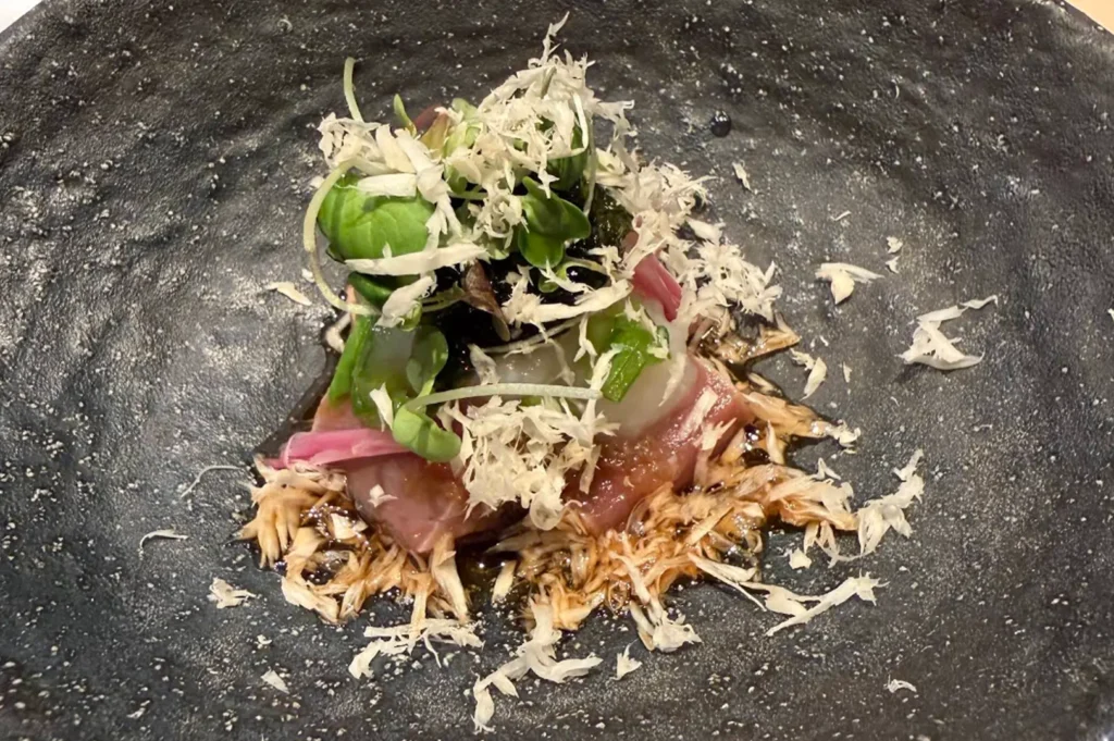 A small, gourmet appetizer is served in a dark, textured bowl, featuring a piece of high-quality fish topped with a delicate mound of microgreens, seaweed, and a sprinkle of light-colored, fluffy shavings. The dish is garnished with thin slices of pickled ginger and served in a rich, dark sauce, emphasizing the meticulous plating and focus on both texture and visual presentation characteristic of fine dining omakase.