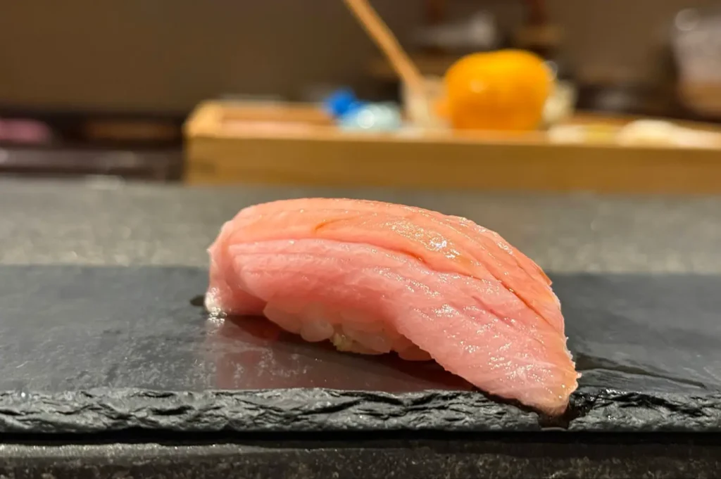 A single, pristine piece of fatty tuna nigiri, with its delicate, marbled pink texture clearly visible, rests centrally on a dark, matte slate surface, with the blurred background of a professional sushi counter visible behind it.