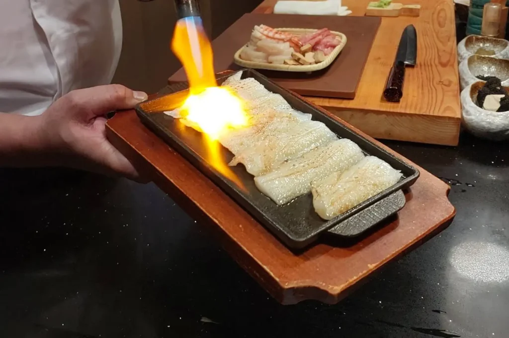 A chef’s hands hold a wooden board supporting a small rectangular cast-iron skillet, using a hand-held blowtorch to sear a row of white fish fillets with a bright, fiery orange flame. In the background on a raised wooden prep counter, various raw ingredients including shrimp, scallops, and sliced fish are neatly arranged on a small bamboo tray alongside a sharp Japanese kitchen knife and small seasoning bowls.