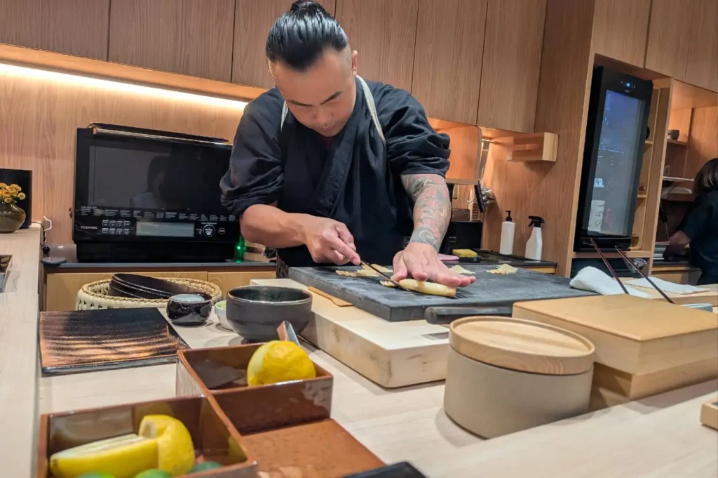 A sushi chef with tattooed arms stands behind a light wood omakase counter, carefully slicing fresh fish on a dark stone cutting board surrounded by prep bowls, citrus fruits, and kitchen equipment.