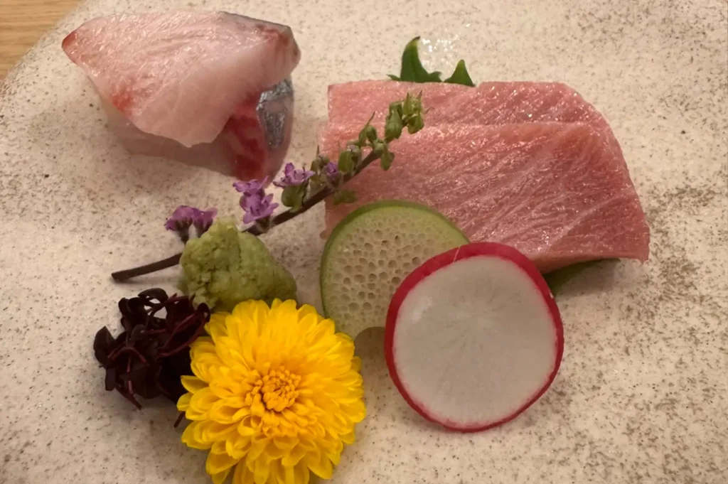 Thickly sliced pieces of fresh sashimi, including marbled fatty tuna and white fish, are arranged on a speckled off-white plate. The dish is elegantly garnished with a bright yellow chrysanthemum flower, a thin slice of red radish, a piece of green root, freshly grated wasabi, microgreens, and a small sprig of purple flowers.