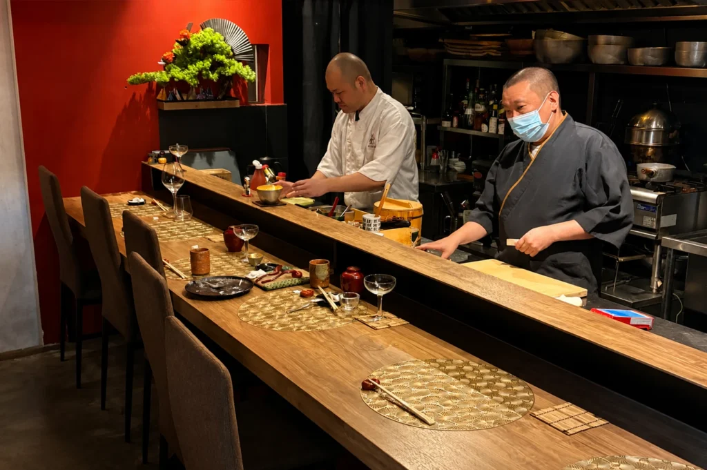 A medium shot capturing two sushi chefs in their professional kitchen, one in a white uniform and the other in a black robe with a blue face mask. They stand behind a long, polished wooden sushi counter in a restaurant with red walls and a decorative bonsai, preparing ingredients with precision for an exclusive dining experience.