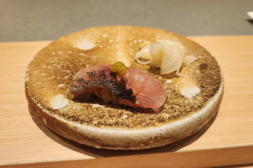 A single piece of nigiri sushi, featuring a slice of seared fish topped with a dollop of green garnish, is presented on a round, textured, earth-toned ceramic plate. Beside the sushi, a small mound of thinly sliced, pale pickled ginger rests on the plate, which sits atop a light-colored wooden table.