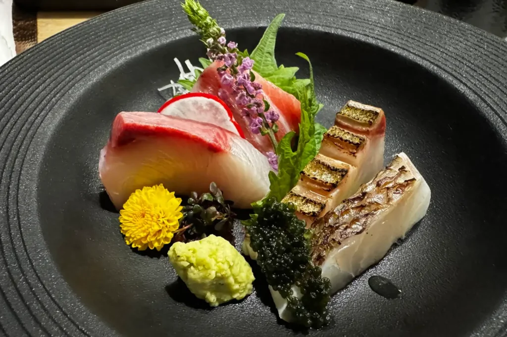 A close-up view of an omakase sashimi platter served in a textured black ceramic bowl, featuring thick slices of fresh fatty tuna (otoro), sea bream, and lightly seared white fish with charred skin, garnished with a bright yellow edible flower, purple shiso blossoms, sea grapes, a shiso leaf, radish slice, and a dollop of fresh wasabi.