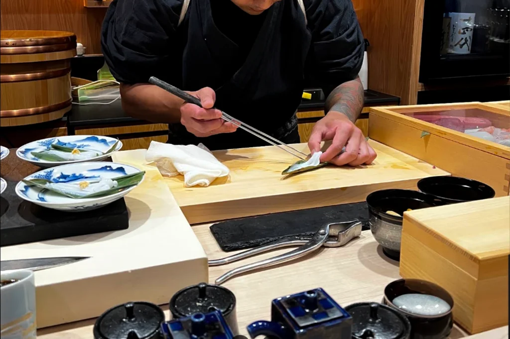 A sushi chef dressed in a black uniform is captured mid-action, using long metal chopsticks to carefully prepare a delicate ingredient on a light wooden cutting board, with small ceramic bowls and serving plates arranged nearby.