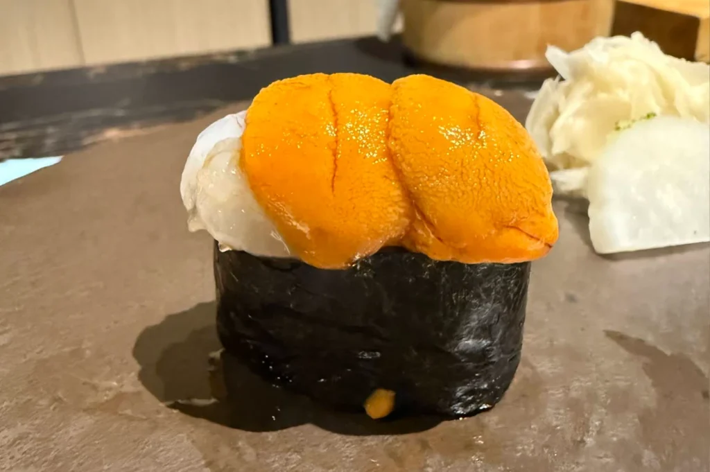 A tight close-up features a piece of premium gunkan maki sushi presented on a rustic, dark brown ceramic serving block. The sushi consists of a crisp sheet of dark nori seaweed wrapped tightly around a base of rice, overflowing with a generous, decadent mound of bright, creamy orange sea urchin (uni) nestled next to a piece of sweet, translucent botan ebi (botan shrimp). In the blurred background on the right, a side of pale pickled ginger and a slice of white radish sit on the counter.