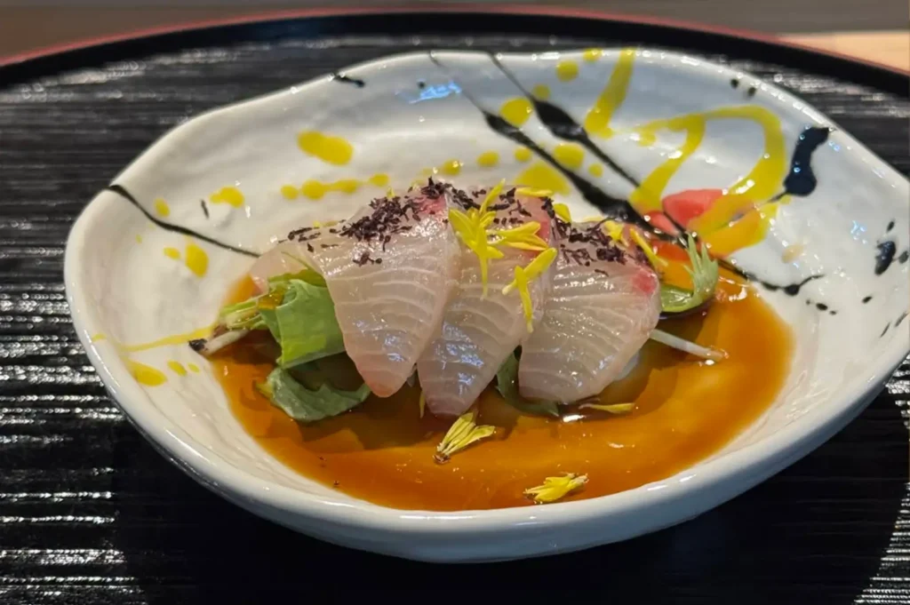 A close-up shot displays a delicate seafood dish served in an organic, white ceramic bowl with black and yellow abstract brushstrokes. Several thinly sliced, translucent pieces of white fish sashimi are elegantly layered over a small bed of fresh green leaves, garnished with vibrant yellow edible petals and a sprinkling of dark purple herbs. The entire arrangement sits in a shallow pool of savory, amber-colored soy-based sauce, all resting on a larger textured black plate.