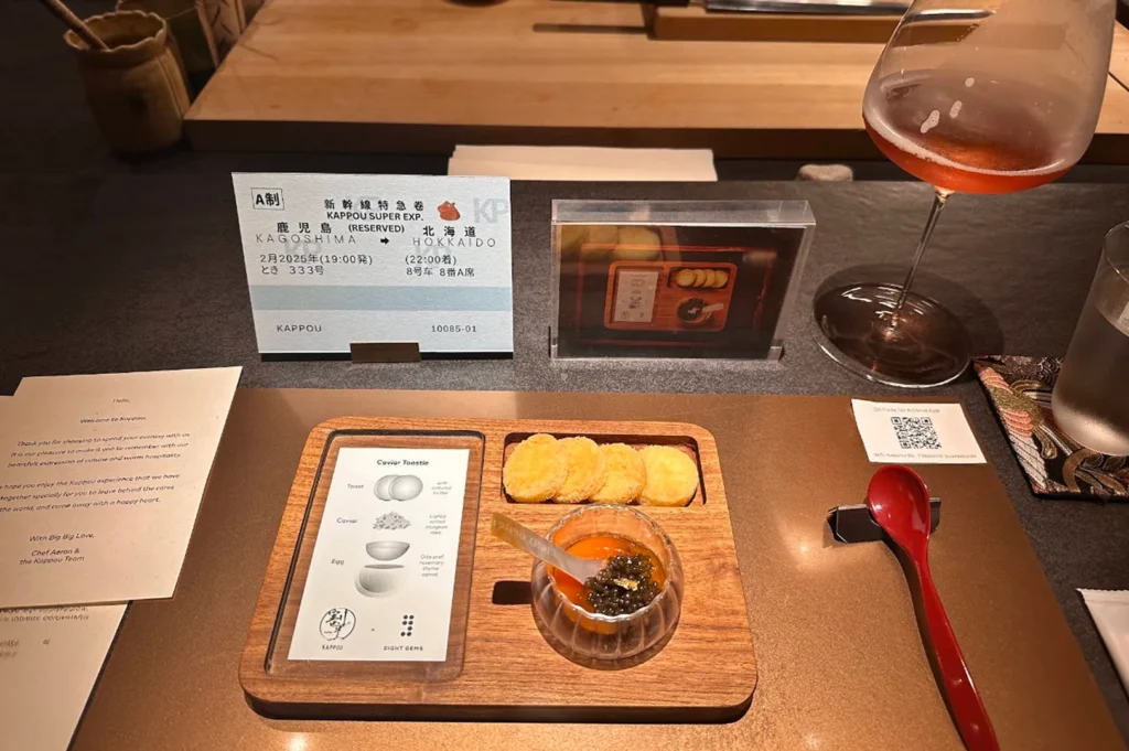 A sophisticated, personalized dining place setting on a metallic bronze placemat at a Kappou counter. The center features a custom wooden tray displaying a "Caviar Toastie" set: four small, golden-brown circular toasts next to a glass dish of bright orange egg yolk topped with a mound of black sturgeon caviar. Accompanying the dish are a personalized welcome card, a novelty Shinkansen train ticket marked "Kappou Super Exp.," a red spoon on a ceramic rest, a framed photo, and a glass containing a reddish-amber beverage.