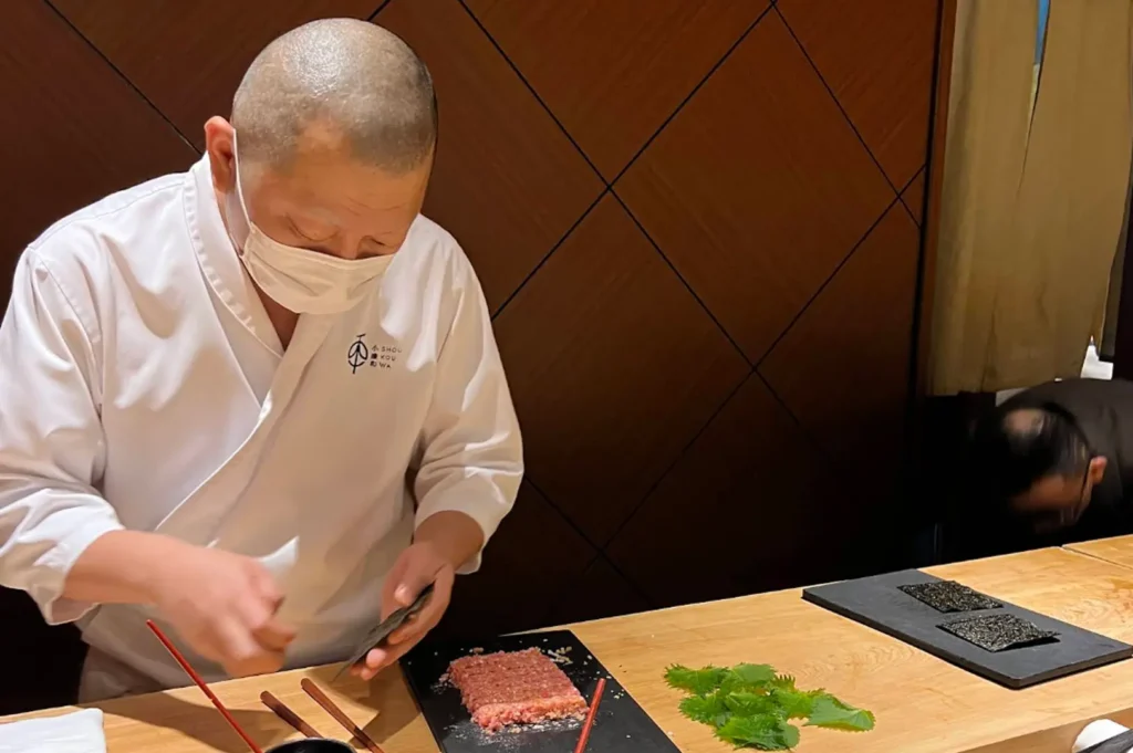 A sushi chef wearing a white uniform and a face mask is captured mid-preparation at a wooden counter, holding a small tool while a block of minced raw tuna and fresh green leaves are laid out on a black rectangular serving stone.