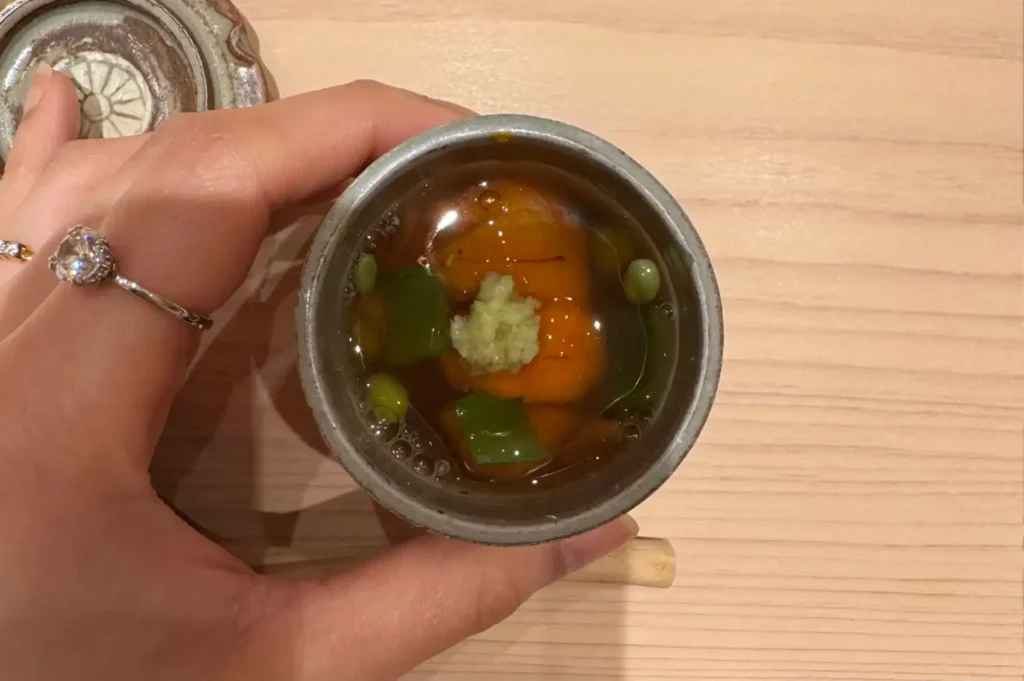 A top-down view of a hand holding a small, rustic ceramic bowl filled with fresh orange sea urchin (uni), green garnishes, and a dollop of grated wasabi suspended in a delicate, clear dashi jelly.