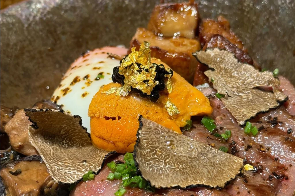 An extreme close-up shot reveals a luxurious gourmet rice bowl filled with decadent layers of high-end ingredients. At the center sits a bright orange mound of fresh uni topped with glossy black caviar and flakes of pure gold leaf. Surrounding the uni are thin, beautifully marbled slices of medium-rare Wagyu beef, cooked mushrooms, cubes of seared foie gras, a soft-poached egg with seasoned edges, and several large, thin shavings of aromatic black truffle, all garnished with finely chopped green chives.