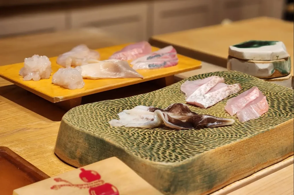 Various delicate cuts of raw fish, shell, and seafood sashimi are artfully arranged across raised wooden and patterned green ceramic serving blocks on a clean wooden counter.