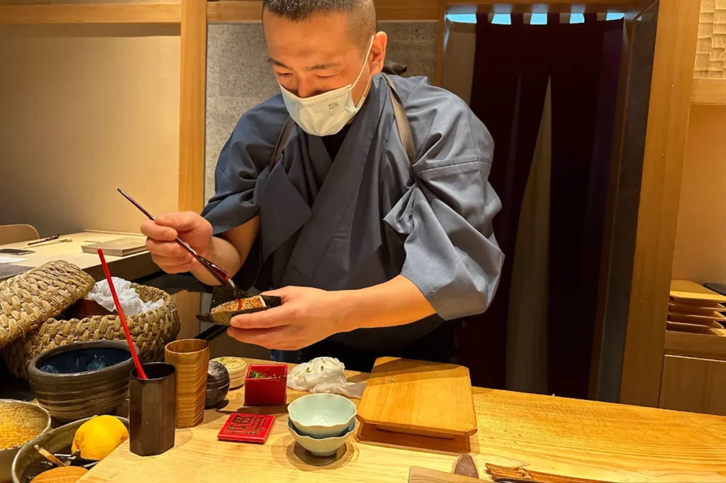 A sushi chef, wearing a mask and a traditional grey kimono-style top, carefully prepares a piece of nigiri behind a light-colored wooden counter. The workspace is neatly organized with small bowls, a wooden serving board, and various ingredients, showcasing the focused, artisanal approach of the chef as he assembles a fresh piece of sushi for the diner.