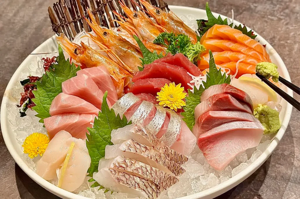 A circular white plate is piled with a variety of fresh sashimi including slices of tuna, salmon, and seared fish arranged over a bed of crushed ice, garnished with green shiso leaves and vibrant yellow edible chrysanthemum flowers, while a pair of chopsticks holds a dollop of bright green wasabi to the side.