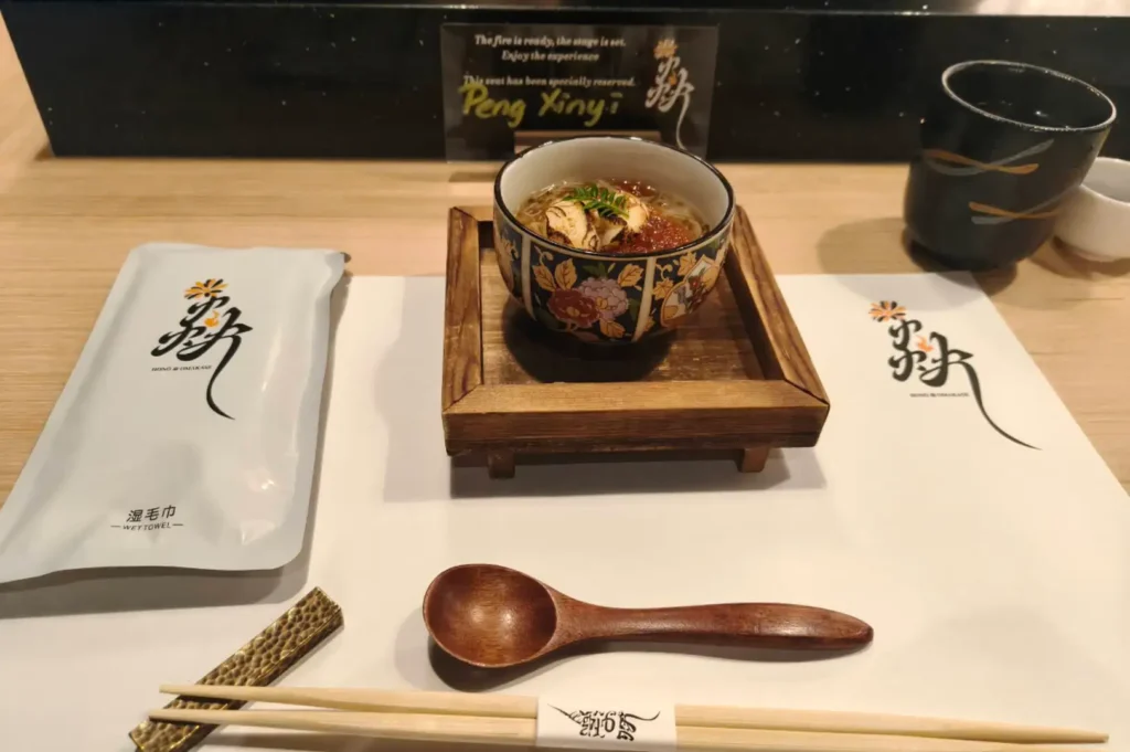 An individual dining setup at an omakase restaurant counter laid out on a clean white placemat. In the center, a raised wooden stand holds a patterned ceramic bowl filled with a warm dish topped with seared ingredients and herbs. Surrounding the bowl are a smooth wooden spoon, a pair of wooden chopsticks resting on a textured brass holder, a packaged wet towel with Chinese and English text, a dark ceramic tea cup, and a small personalized sign in the background reading "Peng Xinyi" with a welcoming message.