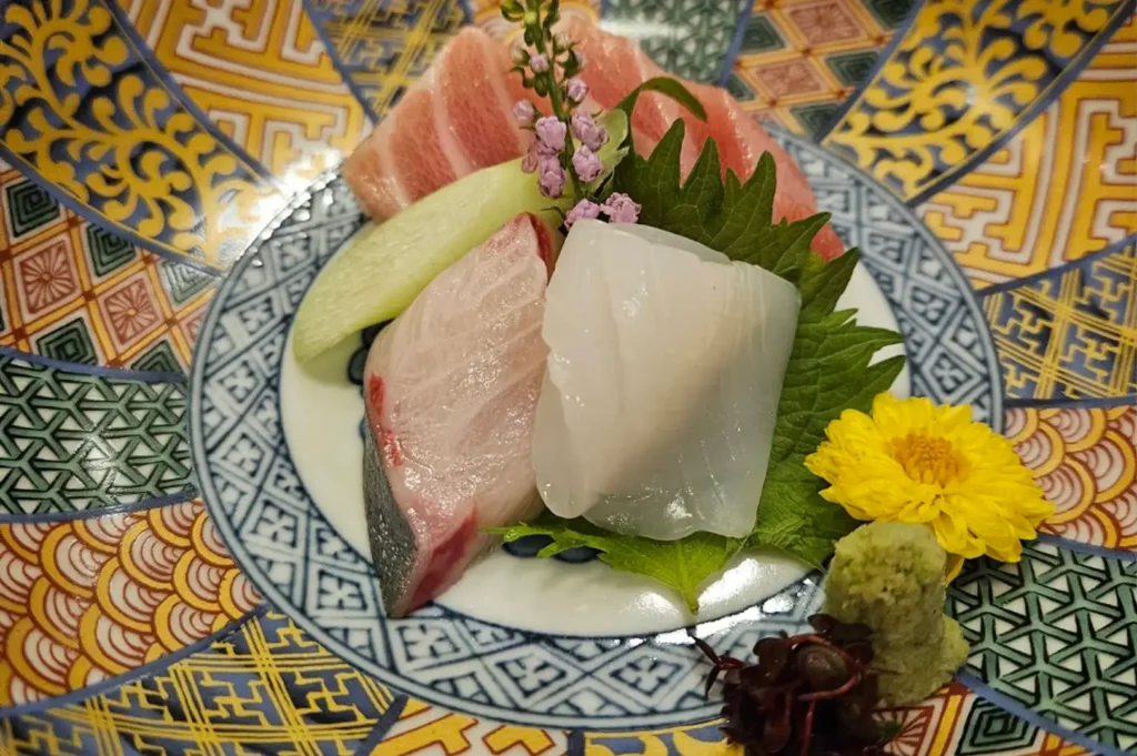 An assortment of raw fish and squid sashimi is artfully presented on a green shiso leaf inside a ceramic bowl featuring intricate traditional Japanese geometric patterns in yellow, red, and blue. The platter is decorated with a vibrant yellow flower, wasabi, red microgreens, and delicate purple blossoms.