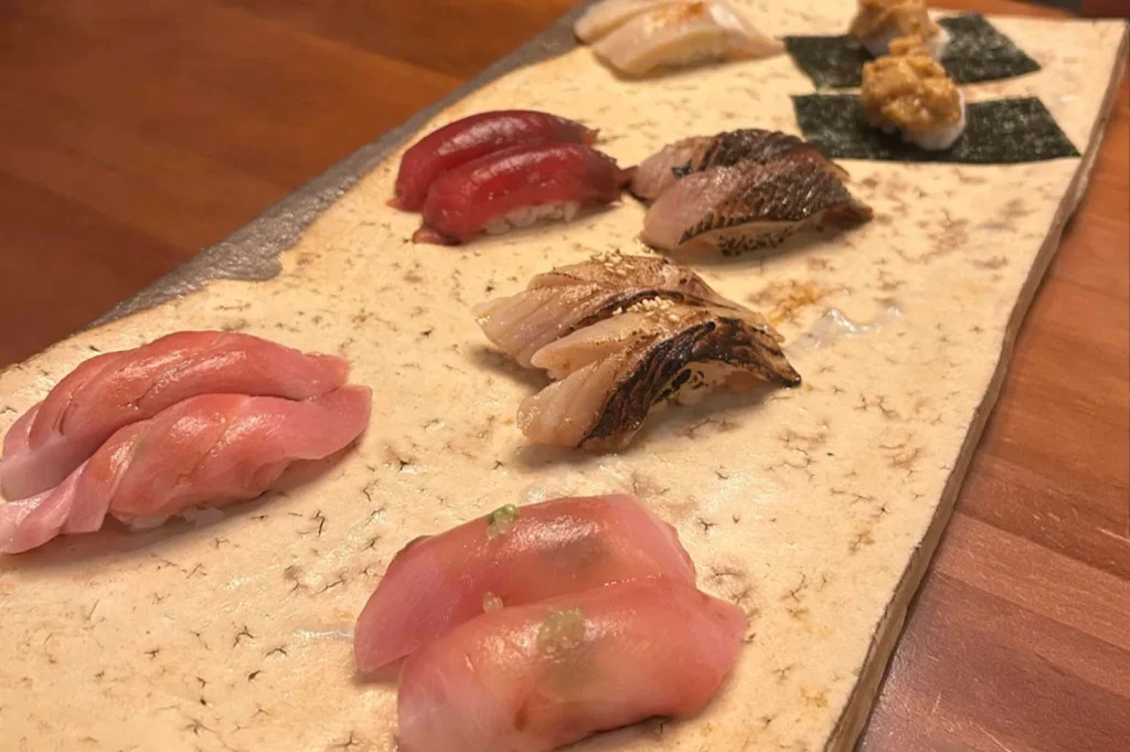 A close-up, high-angle view of a long, light-textured ceramic plate featuring several pieces of expertly prepared nigiri sushi. The selection includes vibrant red tuna, seared white fish with char marks, and pieces topped with sea urchin on nori, all served on a dark wooden table that emphasizes the high quality and craftsmanship of the meal.