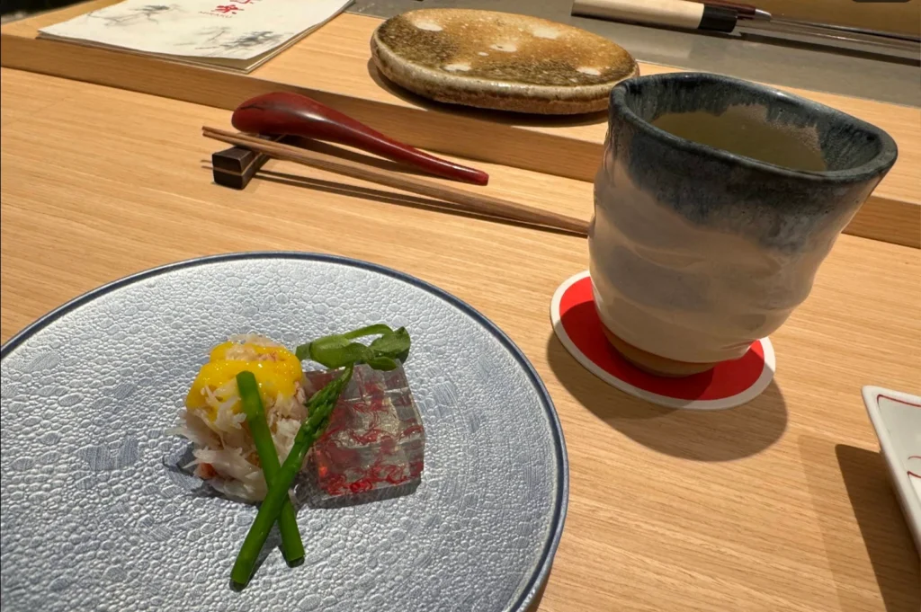A beautifully arranged appetizer sits on a textured, grey-blue plate, consisting of a delicate mound of shredded crab meat topped with a bright yellow sauce and fresh green asparagus stalks, next to a translucent, gelatinous cube containing colorful red ingredients. A textured, white ceramic cup of tea sits on a red coaster nearby, with a red spoon and wooden chopsticks resting on the light wood table in the background.