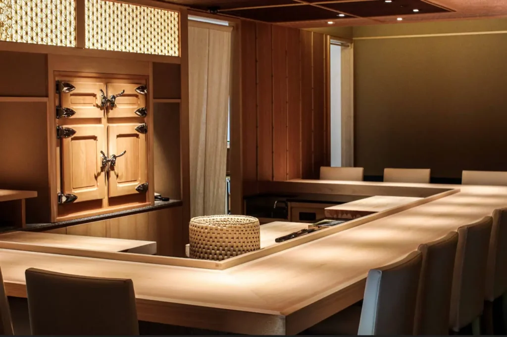 An intimate, minimalist Japanese omakase dining room featuring a polished light wood L-shaped chef's counter lined with cream-colored leather seats, soft overhead spotlighting, a woven bamboo basket, light wood wall paneling, intricate lattice woodwork, and traditional wooden ice-box cabinets in the background.