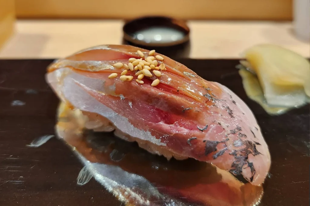 A piece of nigiri sushi topped with a slice of dark, marbled fish and sprinkled with toasted sesame seeds is centered on a black plate, with a hint of pickled ginger blurred in the background.