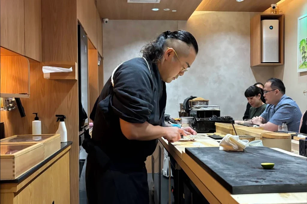A side-angle medium shot captures a male sushi chef with long hair tied back in a neat bun and shaved sides, wearing a dark grey chef's jacket. He stands behind a clean wooden counter, leaning forward and focusing intensely as he uses a traditional knife to slice ingredients on a light wood cutting board. In the foreground, a dark grey stone platter holds a single slice of lime. Across the counter, a few customers are seated, including a man with glasses and a woman with black bangs, watching the chef work within the minimalist, warm-toned restaurant interior.