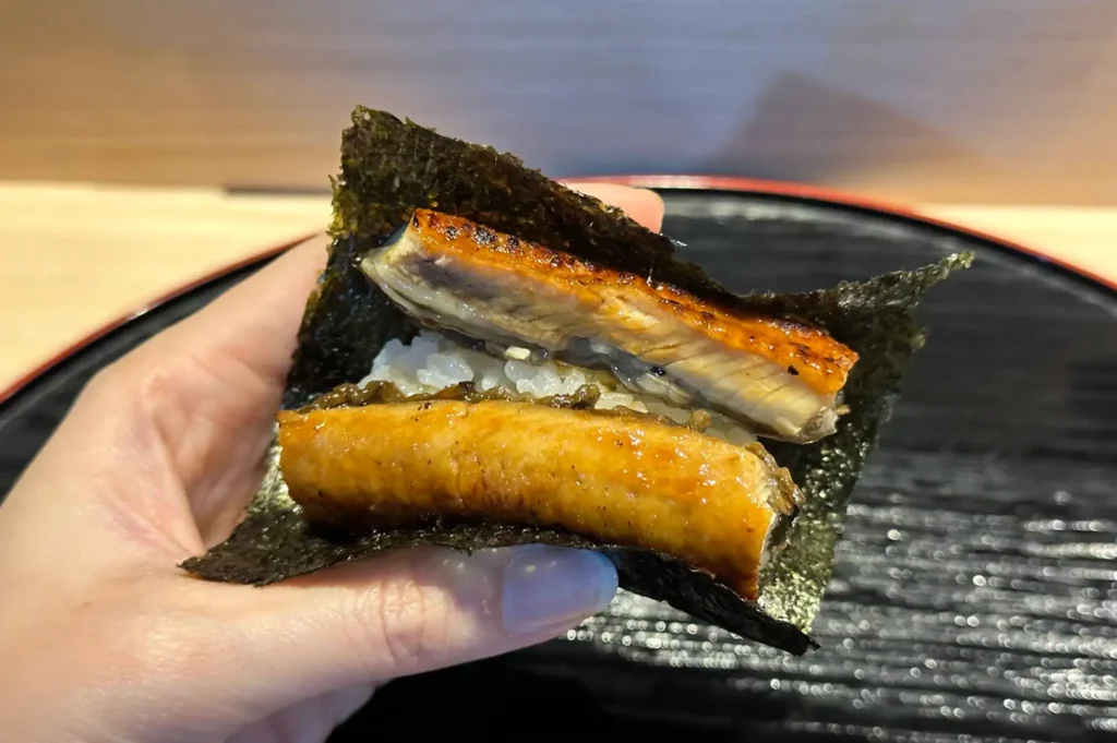 A close-up view focuses on a hand holding a piece of unagi sushi wrapped in a crisp sheet of dark green nori seaweed. Inside the folded seaweed, a bed of seasoned white rice is topped with two thick, glistening pieces of grilled freshwater eel. The fish features a beautifully charred, golden-brown skin coated in a glossy, sweet tare glaze that reflects the light.
