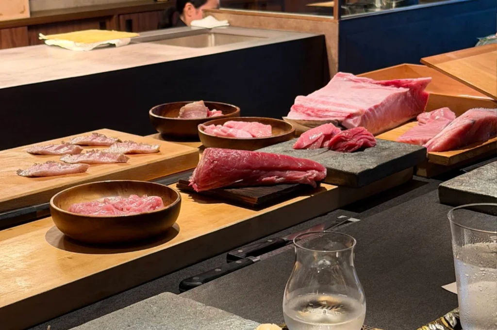 An exquisite display of raw, premium seafood cuts arranged on wooden boards and dark stone slabs along a restaurant counter. The selection includes delicate slices of white fish lightly seared on one side, a dark red block of lean tuna, small wooden bowls filled with minced pink fish, and a massive, beautifully marbled block of pink fatty tuna (otoro) resting on a wooden tray. In the blurred foreground, clear glasses of water sit on the dark counter, emphasizing the fresh food in the background.