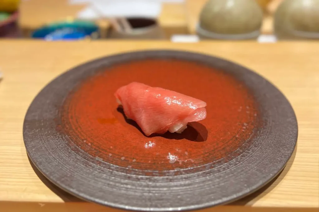A piece of premium tuna nigiri sushi, with a glossy and vibrant red-pink hue, is placed in the center of a rustic, circular brown plate that sits on a light wooden counter.