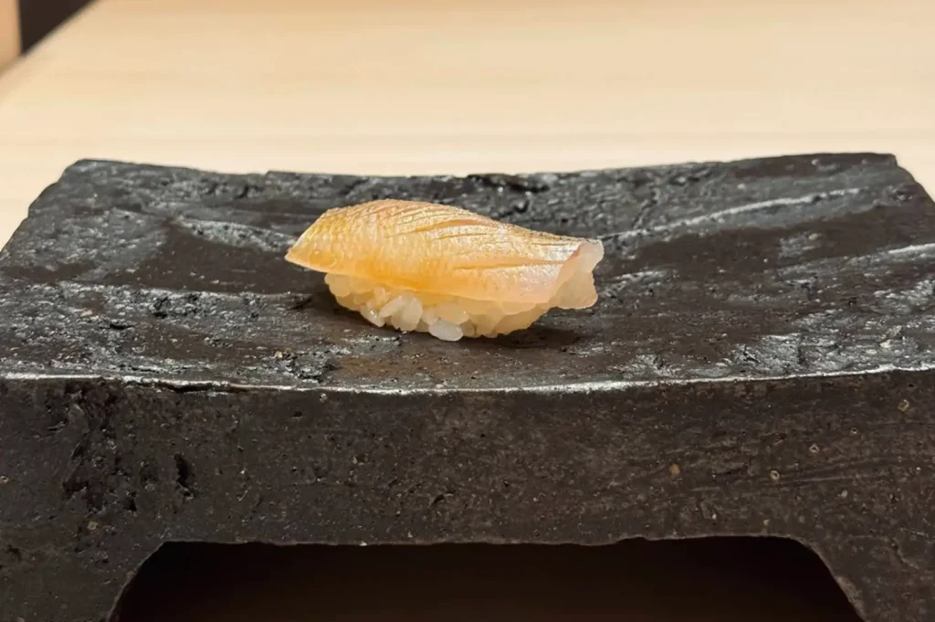 A single piece of delicately brushed white fish nigiri sushi resting on a textured, dark grey elevated stone block plate against a minimalist background.