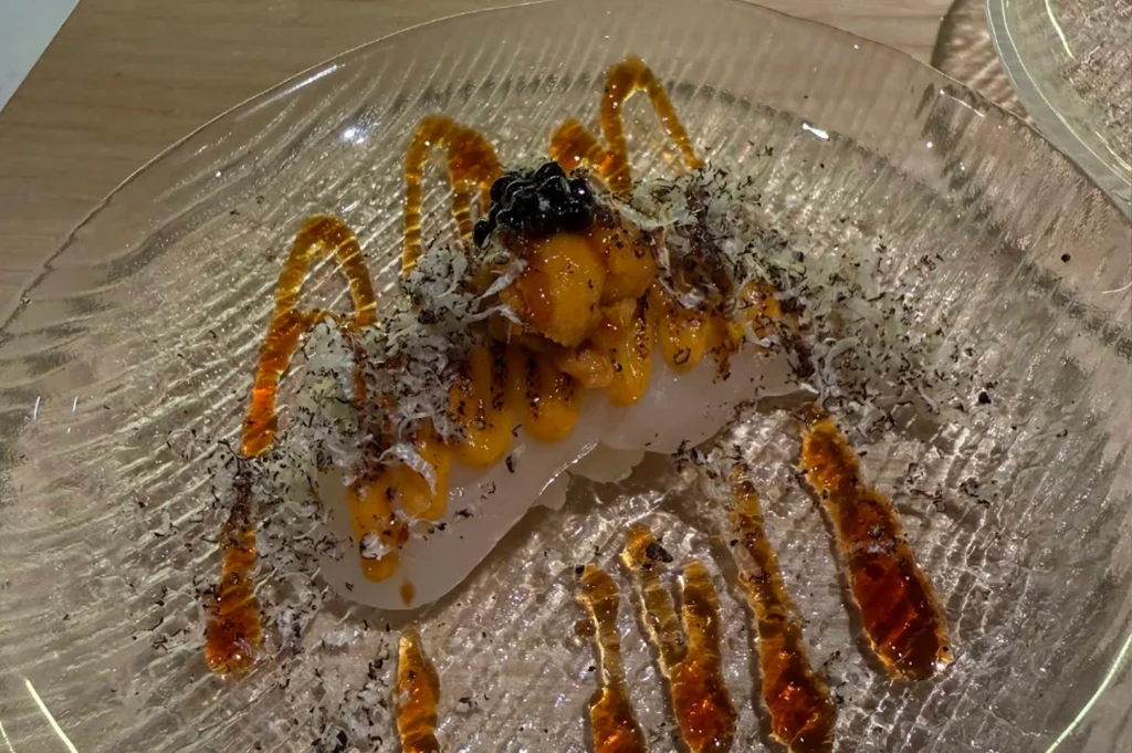 A high-angle close-up showcases a piece of premium nigiri sushi served on an elegant, clear rippled glass plate. The sushi features a base of white seasoned rice topped with a thick slice of translucent white fish, which is neatly layered with bright yellow-orange sea urchin (uni) and a cluster of glossy black caviar. Drizzles of dark, amber-colored soy reduction weave gracefully across the glass surface, while a generous dusting of shaved black truffles is scattered beautifully over and around the dish.