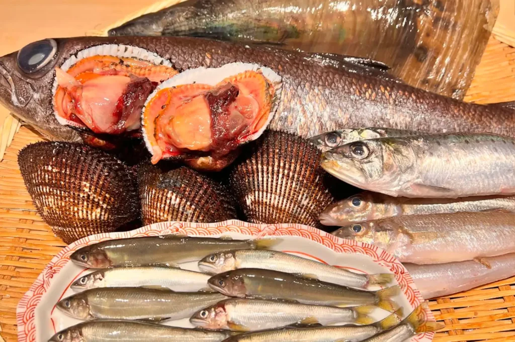 A vibrant, fresh display of raw seafood laid out on a woven bamboo mat features small silvery fish arranged on a red-patterned dish, large black clammed shells with open orange meat, and a large whole dark fish.