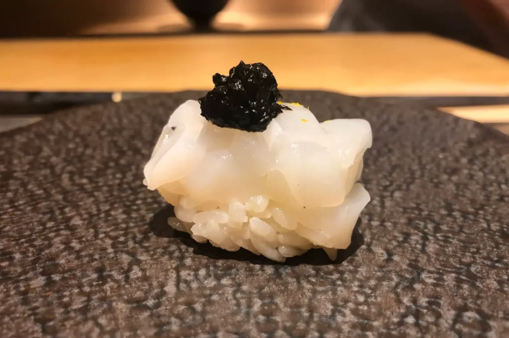 A single piece of nigiri sushi is elegantly presented on a dark, textured plate, featuring a slice of white fish atop perfectly formed sushi rice, crowned with a dollop of glossy, black topping. The simplicity of the plating highlights the quality and freshness of the ingredients, reflecting the precise balance of flavor and aesthetics central to high-end sushi service.