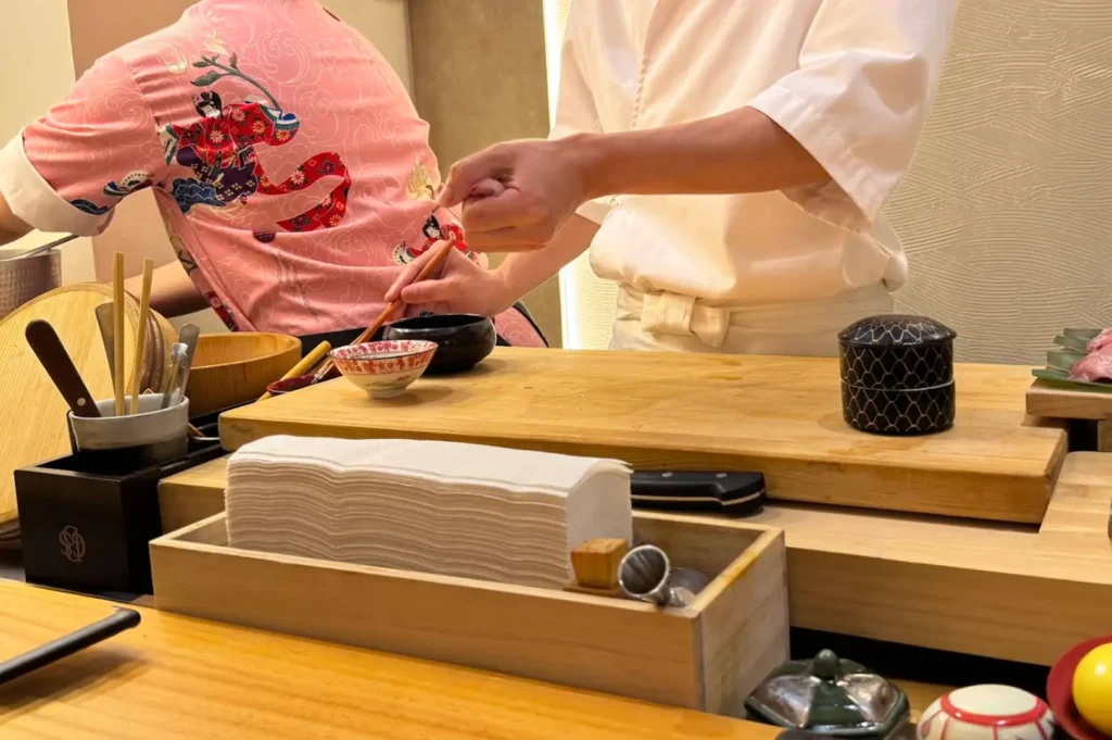 An omakase chef dressed in a crisp white uniform prepares food behind a polished wooden counter, with a pink patterned garment visible on a person to the left, and a neat workspace set up with a stack of white napkins, a small dipping bowl, and other culinary tools.