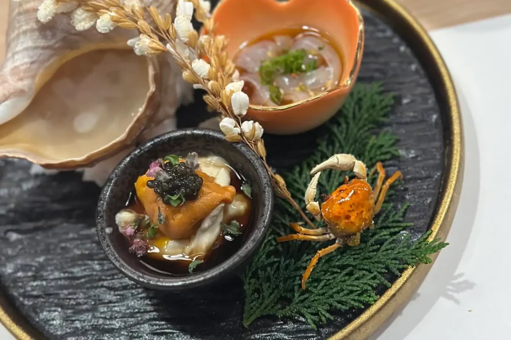 A close-up view of an intricately plated Japanese omakase dish served on a dark, textured round plate with a metallic rim. The arrangement features a large, light-colored conch shell on the upper left, a small black bowl containing creamy sea urchin (uni) topped with dark caviar and delicate purple edible petals, and a small flower-shaped orange bowl filled with translucent seafood garnished with chopped scallions. To the right, a tiny whole crispy fried crab sits on a bed of fresh pine needles, accompanied by a decorative sprig of dried white rice grains resting across the plate.