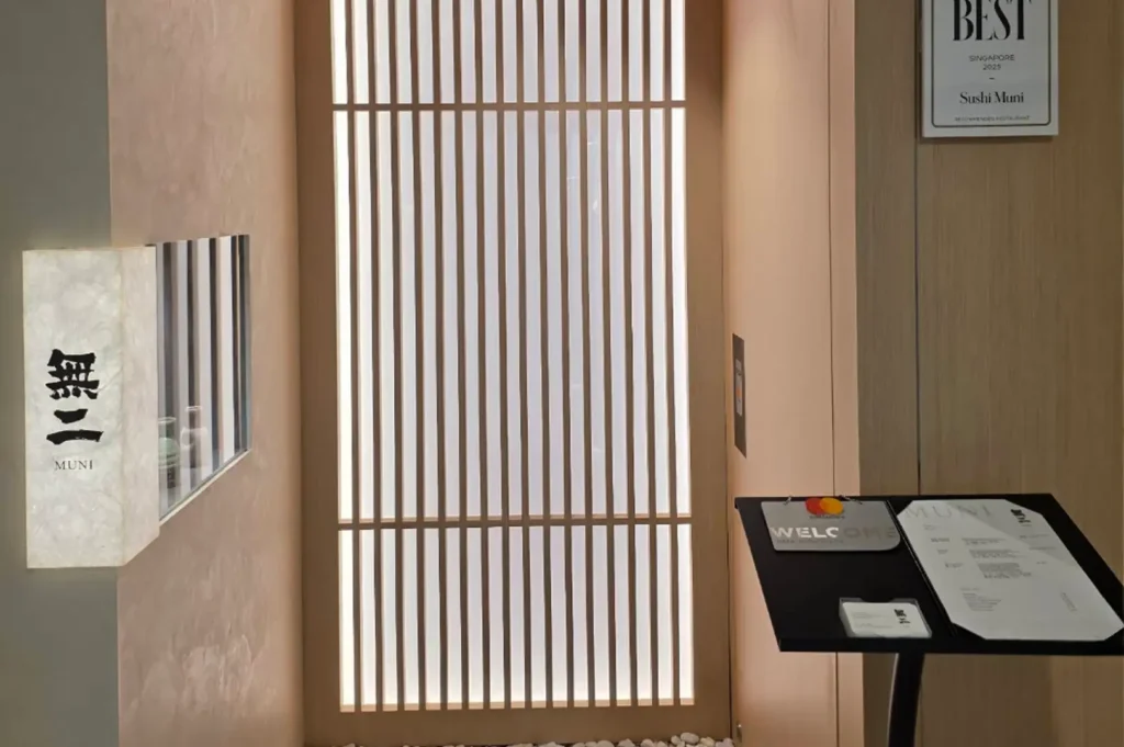 The minimalist entrance of "Sushi Muni" features a traditional Japanese sliding screen door with warm backlighting, a glowing stone sign reading "MUNI" in Japanese kanji, and a menu stand near the doorway.
