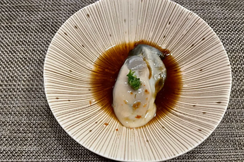 A large, plump fresh raw oyster rests in a shallow pool of dark ponzu sauce inside a textured, light beige ceramic dish with radiating brown lines. A small dab of green garnish and chili flecks top the glistening oyster.