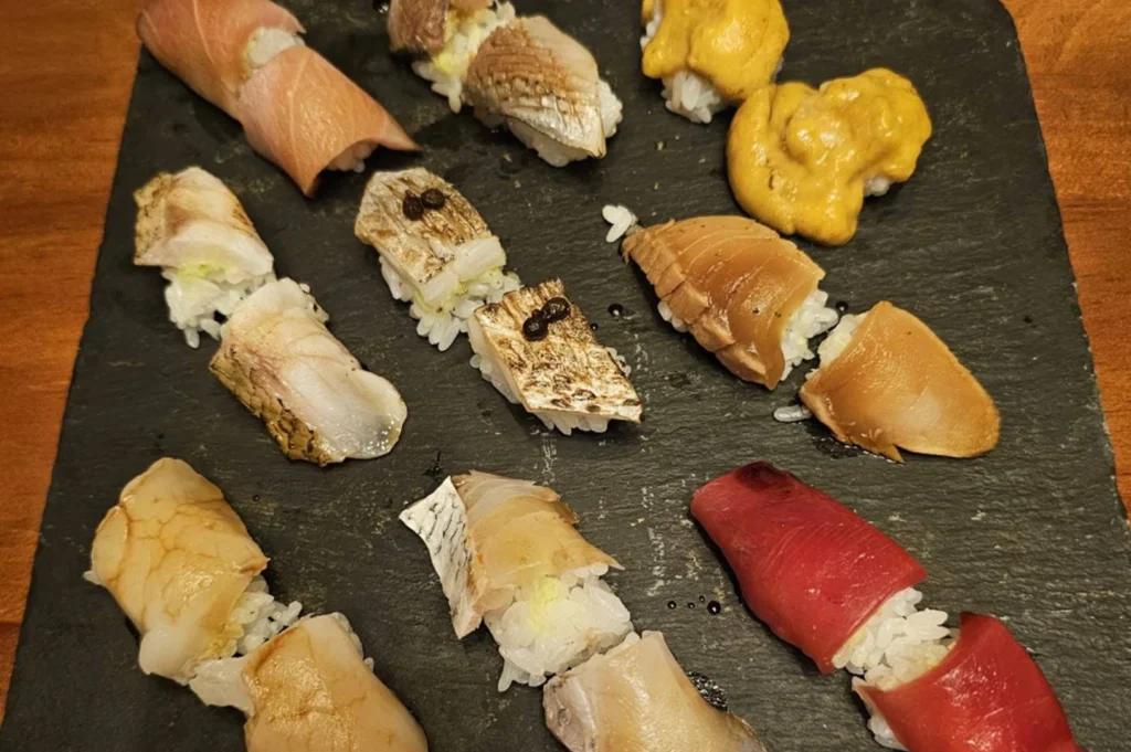 A high-angle, overhead shot of a square, textured black slate plate serving an array of diverse nigiri sushi. The selection includes various cuts of fish, seared toppings, and golden sea urchin, arranged purposefully across the slate surface, showcasing the variety and refined presentation typical of an omakase service.